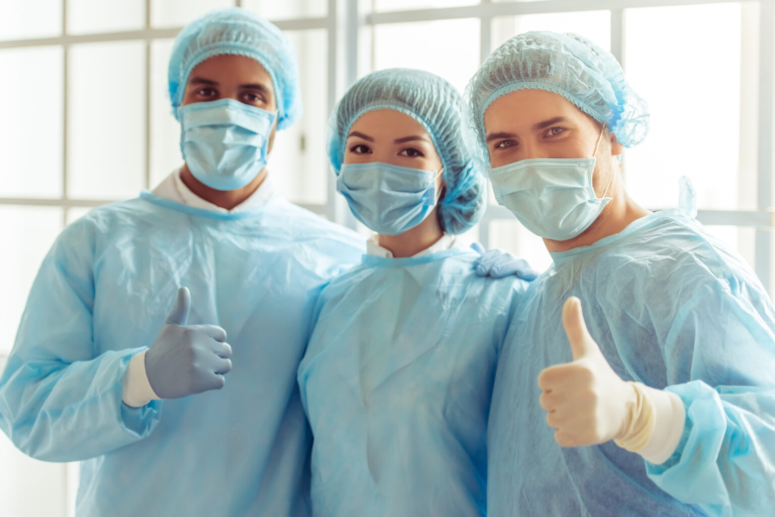 Surgeons of different nationalities and genders in masks and caps are looking at camera. Men are showing Ok sign